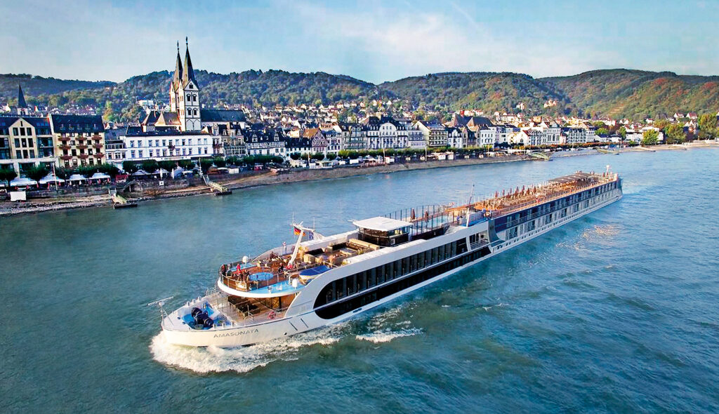 A high-angle aerial view of the AmaSonata river cruise ship sailing on the Rhine River. The ship is white with blue accents and has the name "AmaSonata" written on its side. It is moving from right to left in the image, leaving a white wake behind it. The river is a deep blue color and is surrounded by green hills and vineyards. In the background, there is a picturesque town with a church with a tall steeple. The sky is blue with some white clouds.