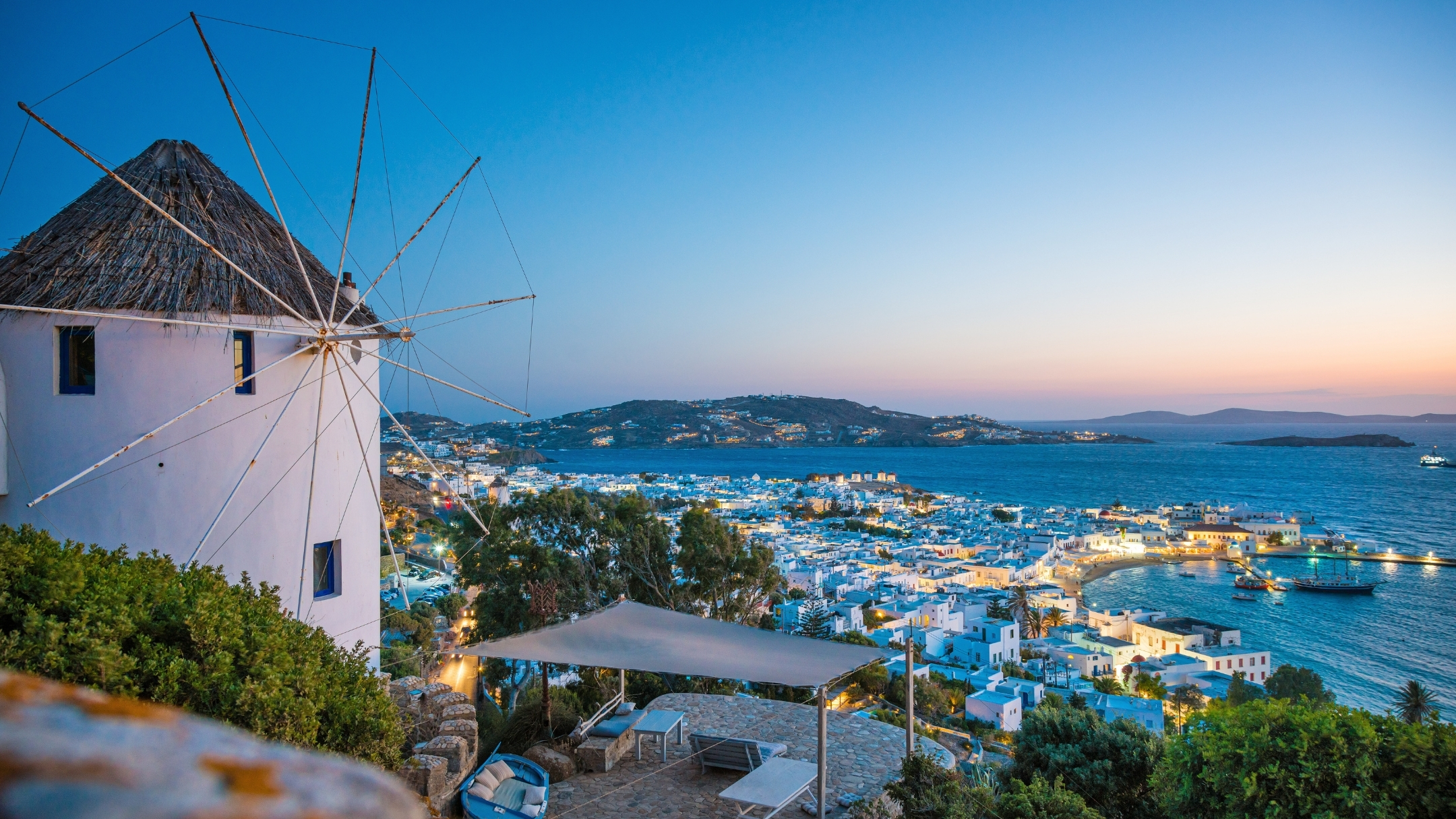 A breathtaking sunset view of Mykonos Town (Chora) on Mykonos Island, Greece. The whitewashed houses and iconic windmills glow in the warm light as the sun dips below the horizon, casting a golden hue over the Aegean Sea.