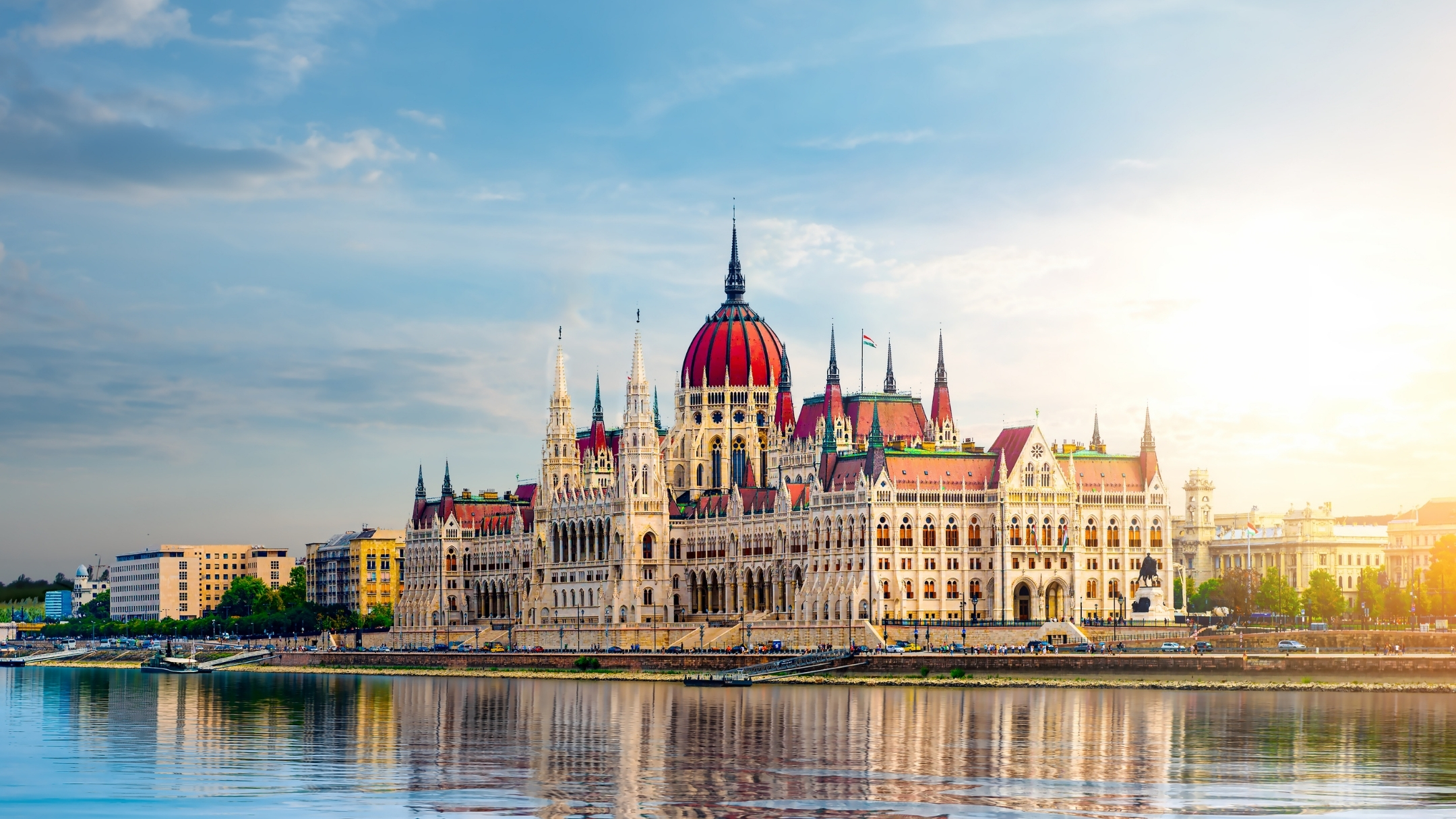 The Hungarian Parliament Building in Budapest, Hungary, seen at sunset. The ornate, neo-Gothic building with its distinctive dome and spires is bathed in warm light from the setting sun. The Danube River is in the foreground, reflecting the building and the sky. A few smaller buildings are visible to the left and right. The sky is a mix of blue with scattered clouds.